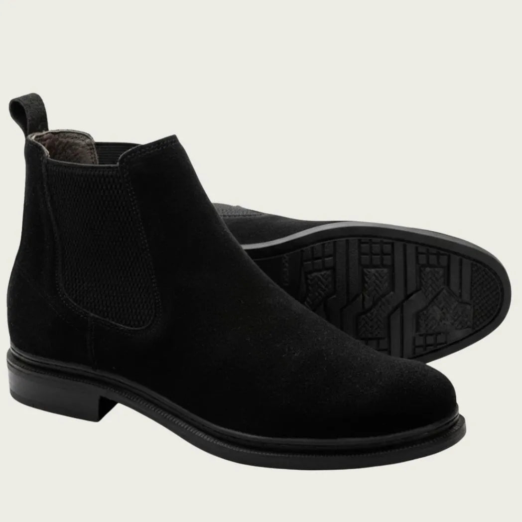 Par de botas Chelsea de Camurça pretas, com um dos solados voltado para a câmera.