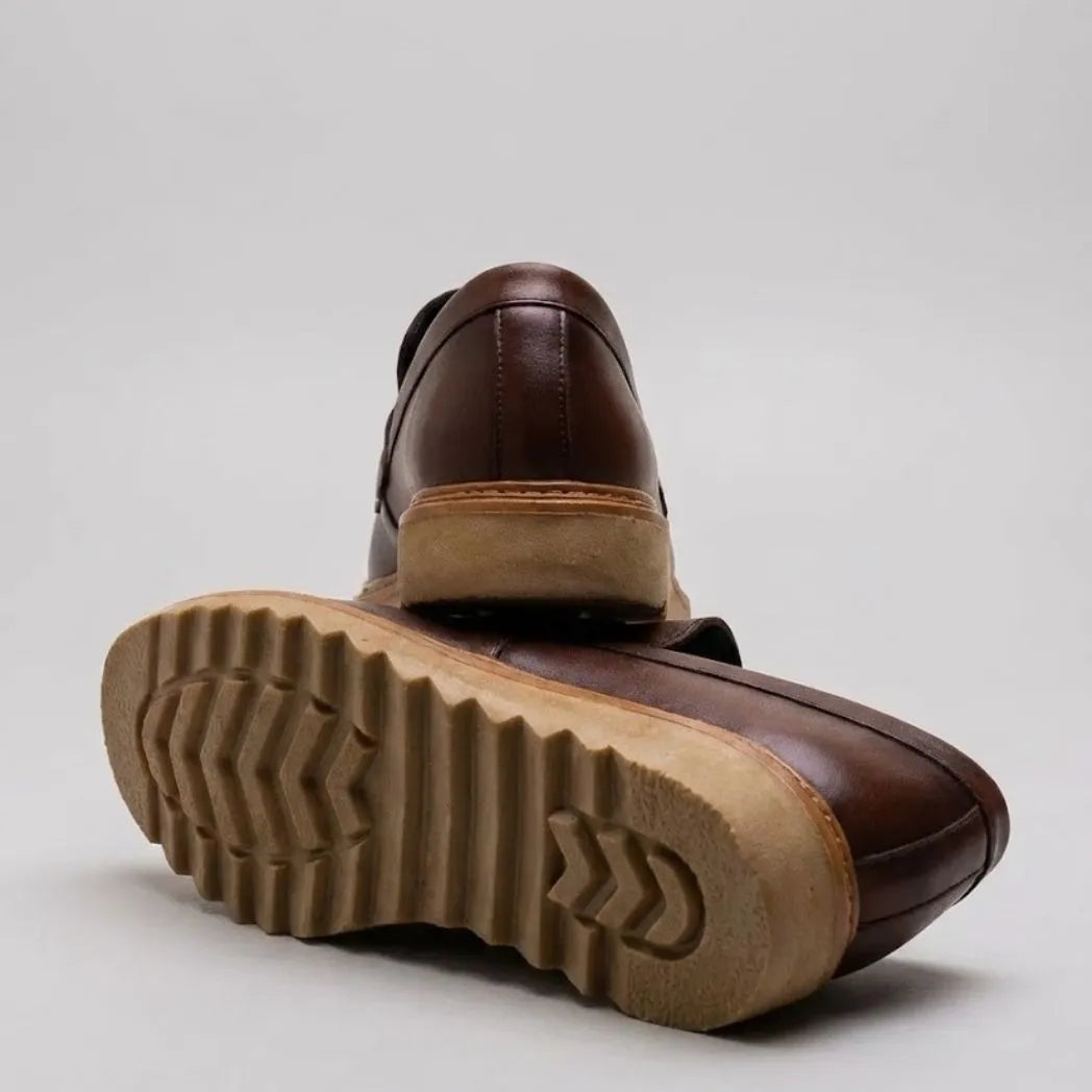 Par do modelo Mocassim Ash Tratorado, na cor marrom, com um dos solados voltado para a câmera.
