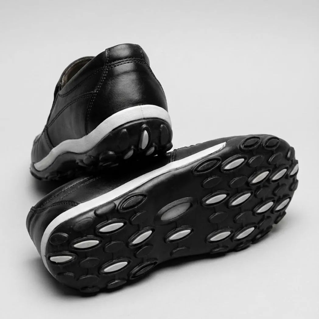 Mocassim Casual Comfort Daytona Drive: par preto com um sapato virado, mostrando a sola preta com detalhes ovais claros.