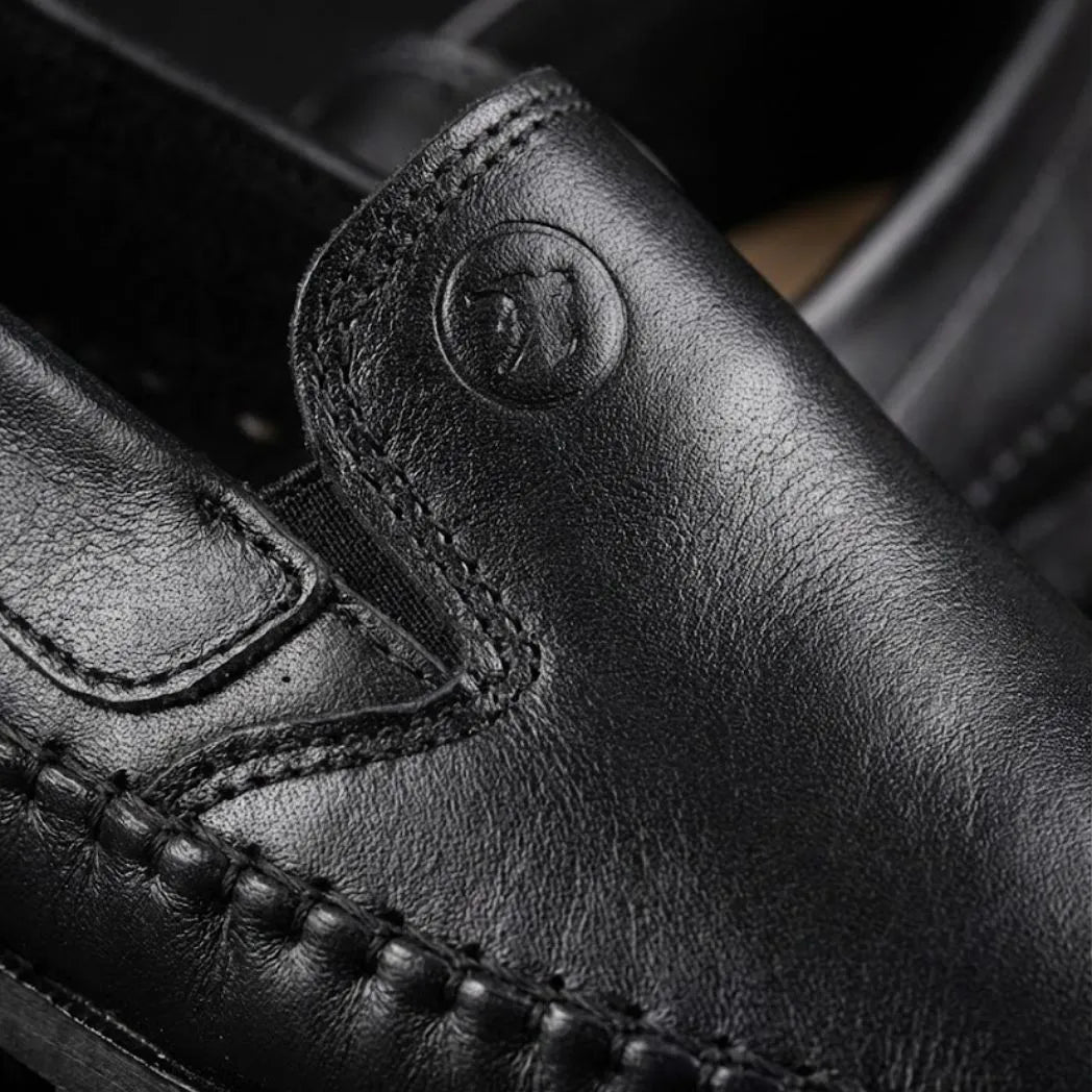 Mocassim Casual Dakota Rust: detalhe da superfície preta, do emblema circular em relevo e da costura.