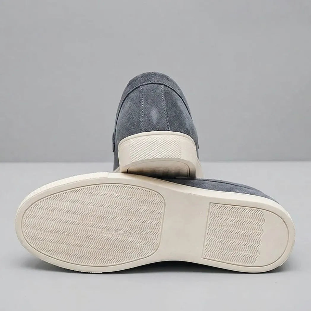 Par do modelo Mocassim Derby de Camurça com Guizos, na cor cinza, com um dos solados voltado para a câmera.