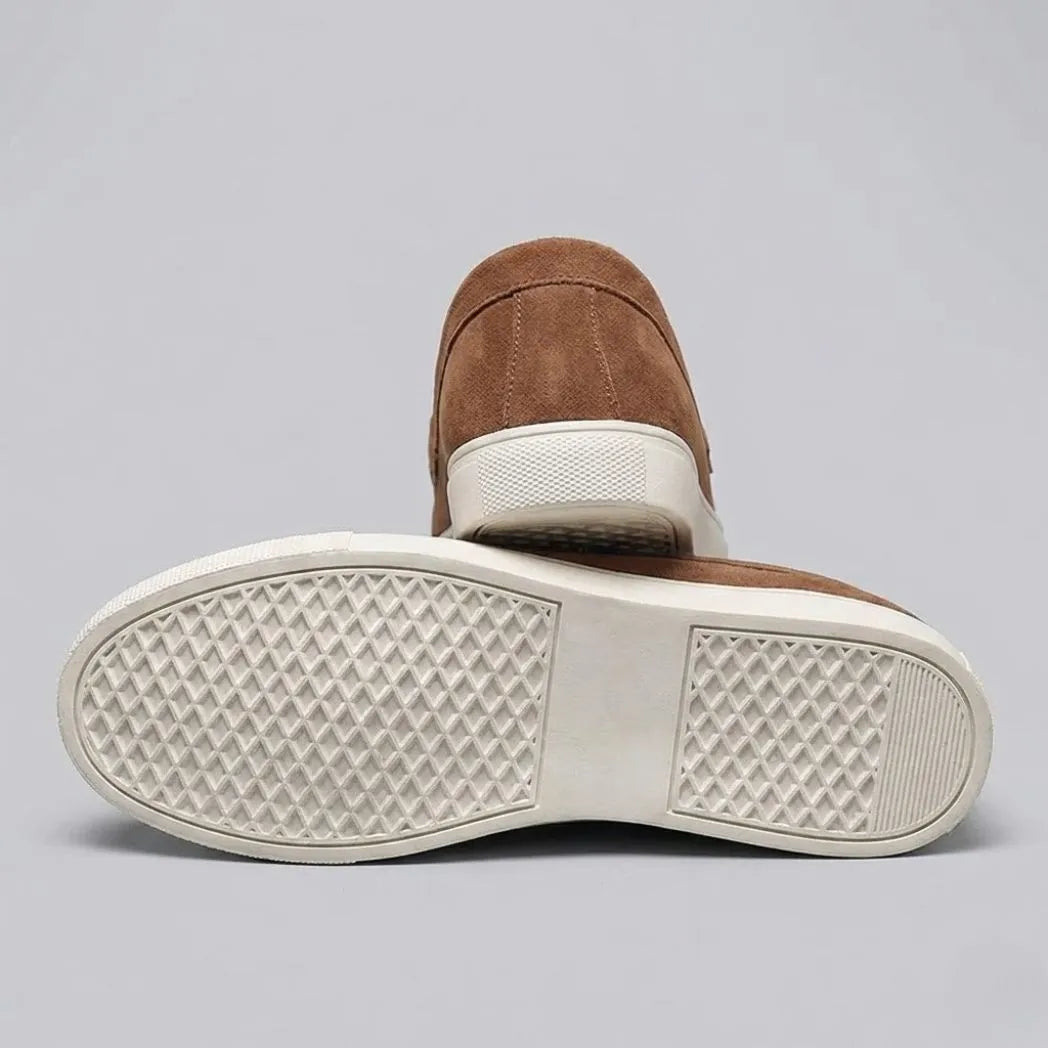 Par do modelo Mocassim Derby de Camurça com Guizos, na cor marrom, com um dos solados voltado para a câmera.