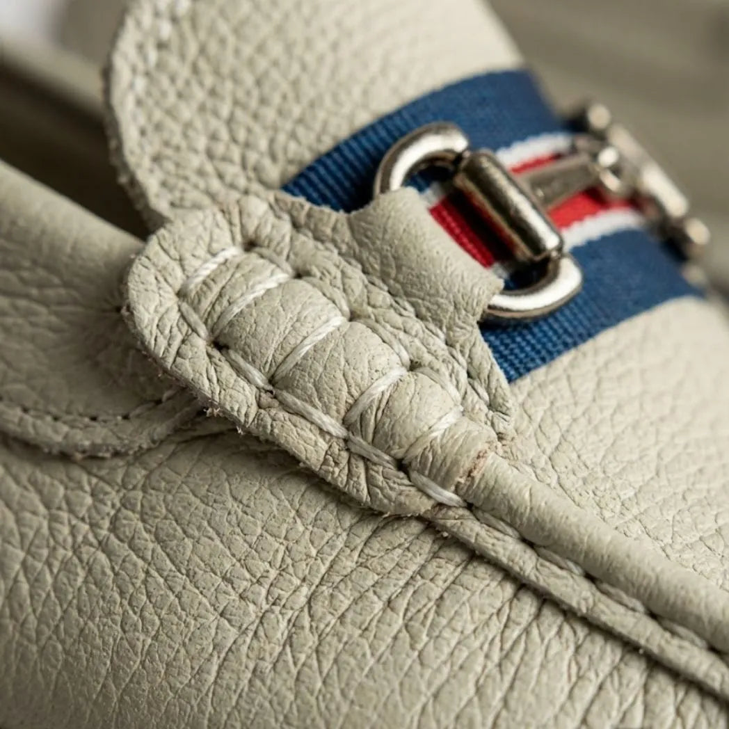 Detalhe da textura e da fivela metálica sobre faixa azul e vermelha do Mocassim Driver Heritage de Couro Branco.
