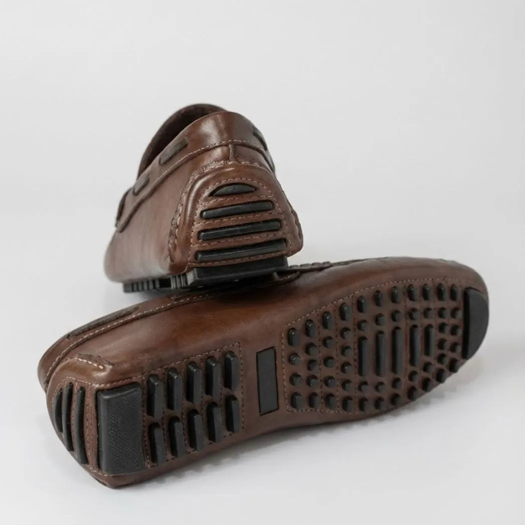 Par do modelo Mocassim Driver Penny de Couro, na cor marrom-escuro, com um dos solados voltado para a câmera.