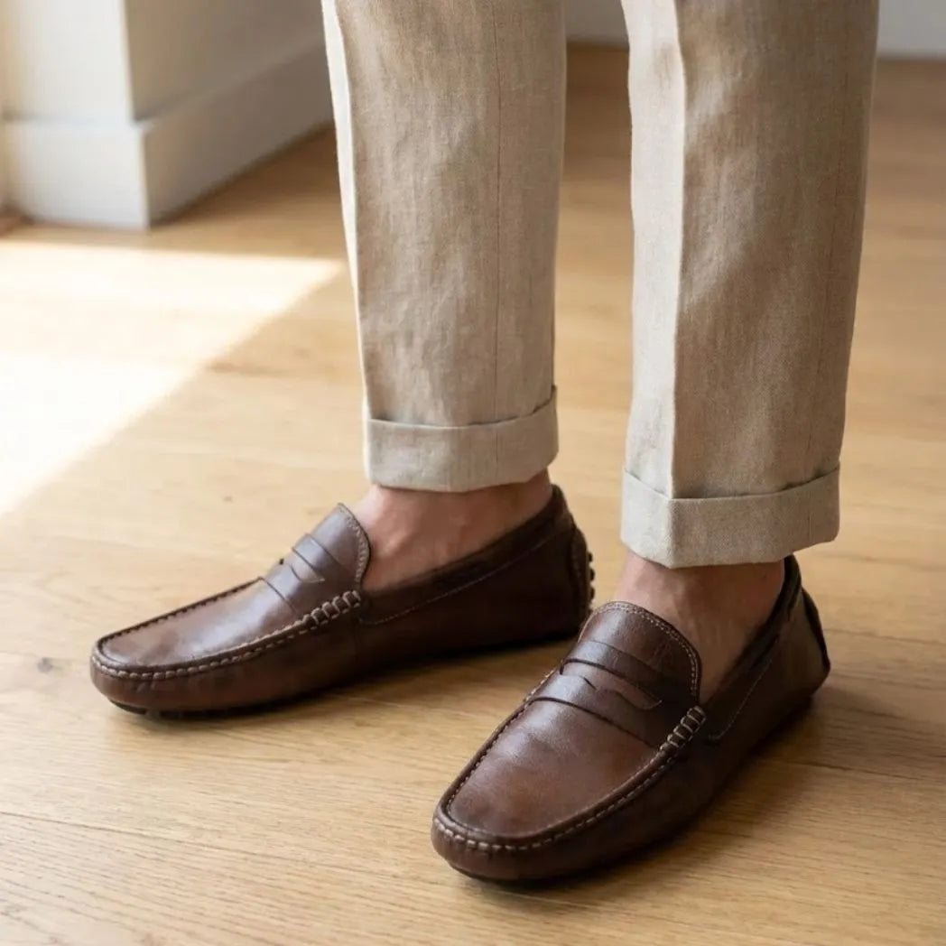Mocassim Driver Penny de Couro na cor marrom-escuro, usado com calça clara de barra dobrada.