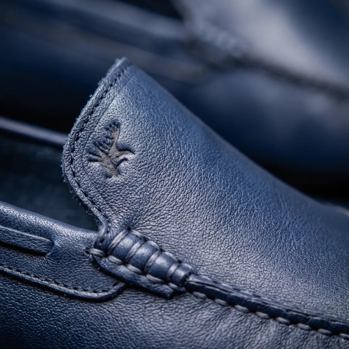 Mocassim Driver Vancouver de Couro Azul: detalhe do couro azul, costura e emblema em baixo relevo, sem metal acrescentado.