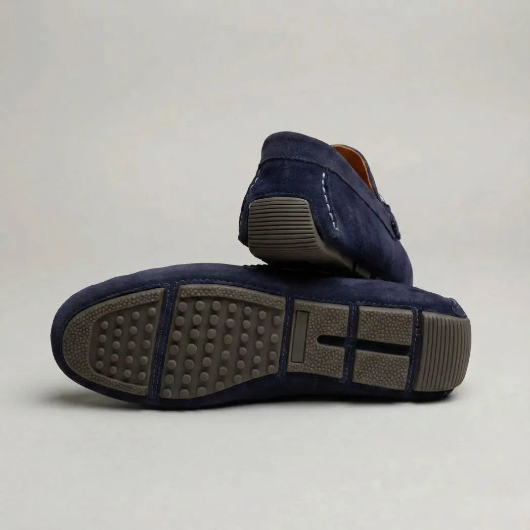 Par do modelo Mocassim Driver de Camurça Capri Dune, na cor azul-escuro, com um dos solados voltado para a câmera.