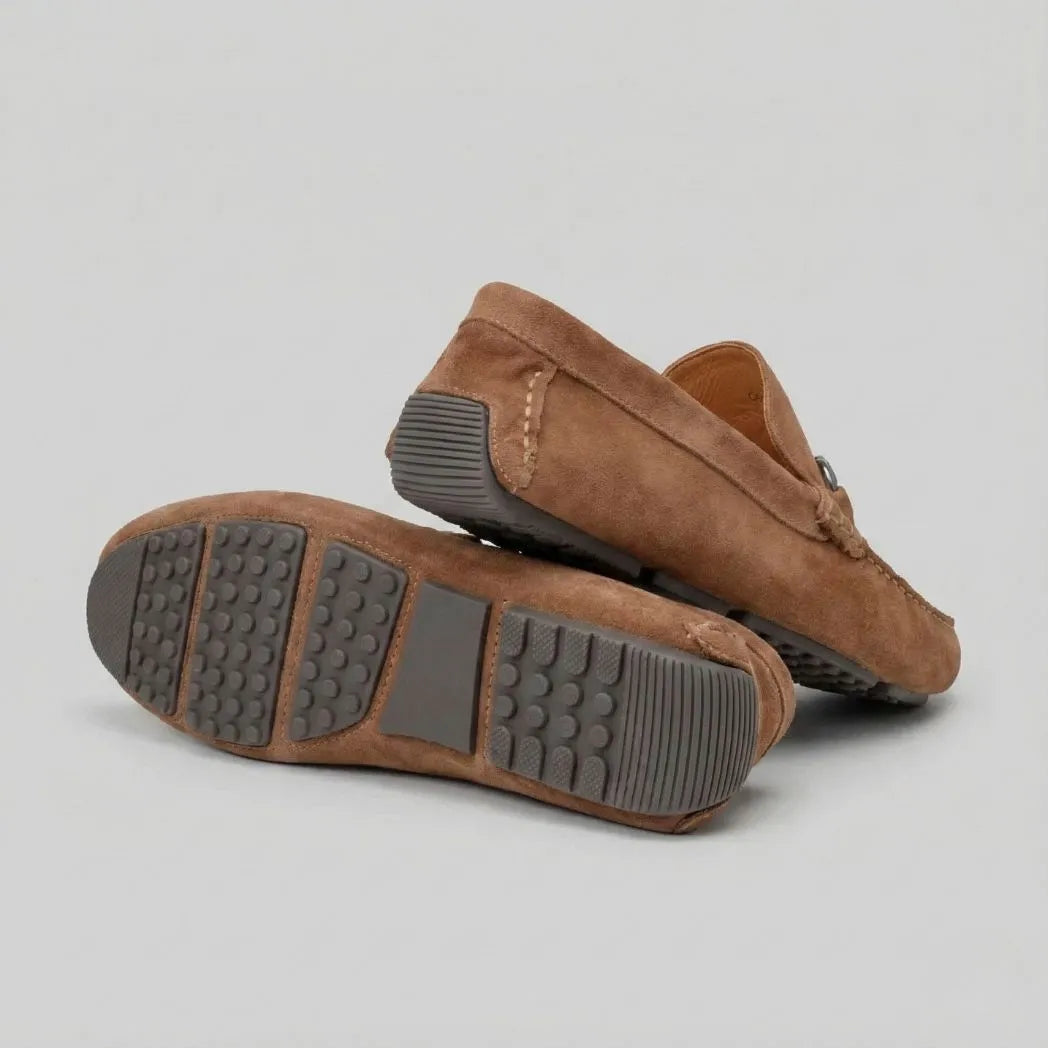 Par do modelo Mocassim Driver de Camurça Capri Dune, na cor marrom, com um dos solados voltado para a câmera.