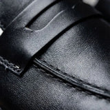 Mocassim Loafer Londres de Couro: detalhe da superfície preta, da faixa frontal e da costura.