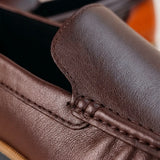 Mocassim Loafer de Couro Contrast Marrom: detalhe do couro marrom e costura escura, com acabamento de contraste ao fundo.