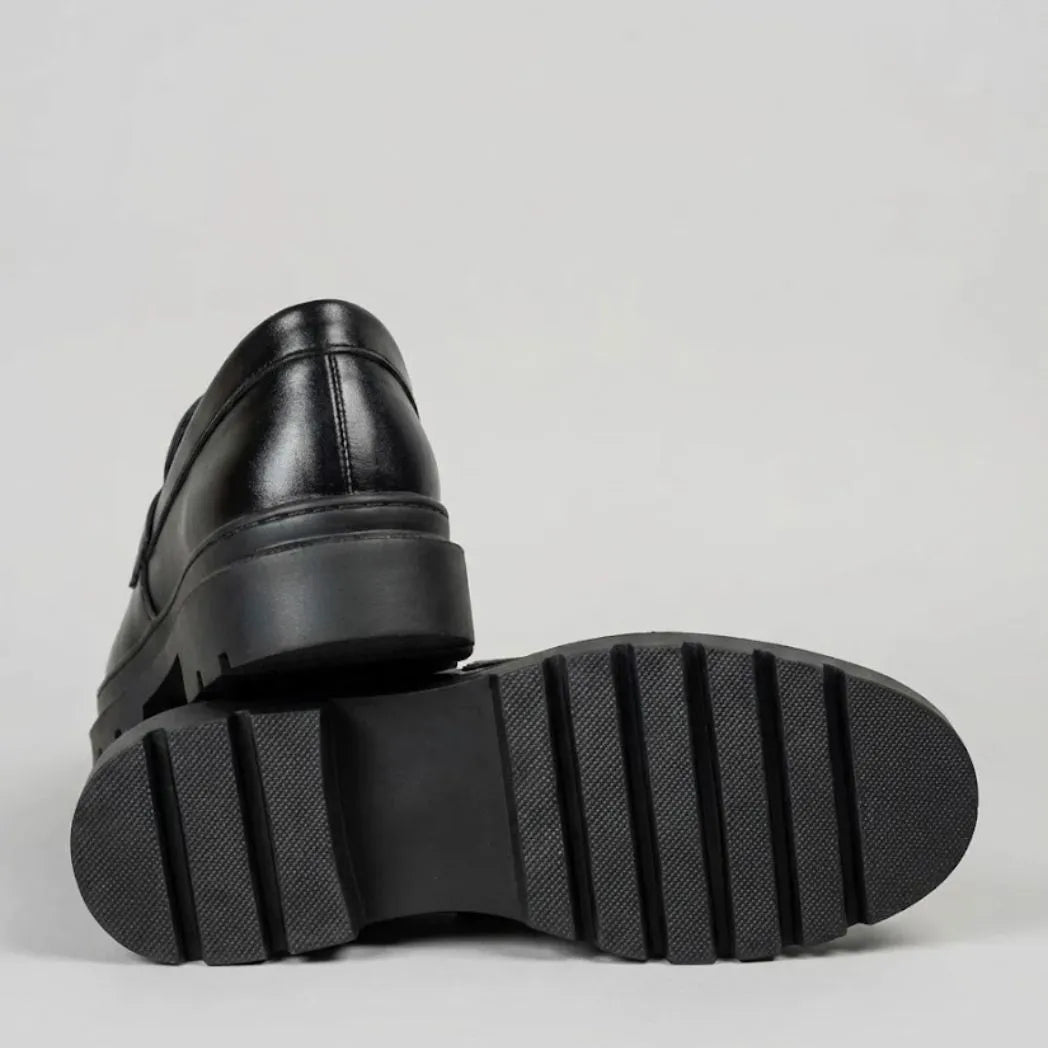 Mocassim Masculino Costurado Piettro: par preto com um sapato virado, mostrando a sola preta com sulcos largos.