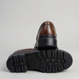 Mocassim Masculino Costurado Piettro: par marrom-escuro com um sapato virado, mostrando a sola preta com sulcos profundos.