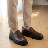 Mocassim Masculino Costurado Piettro: par marrom-escuro usado com calça clara dobrada na barra.