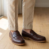 Mocassim Masculino Loafer Atenas: par marrom-escuro com sola bege usado com calça clara dobrada na barra.