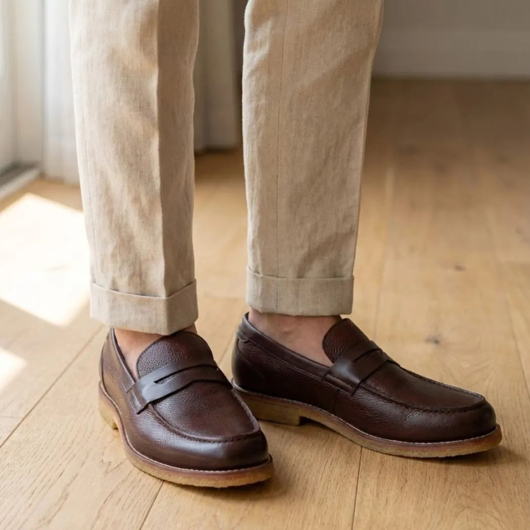 Mocassim Masculino Loafer Atenas: par marrom-escuro com sola bege usado com calça clara dobrada na barra.
