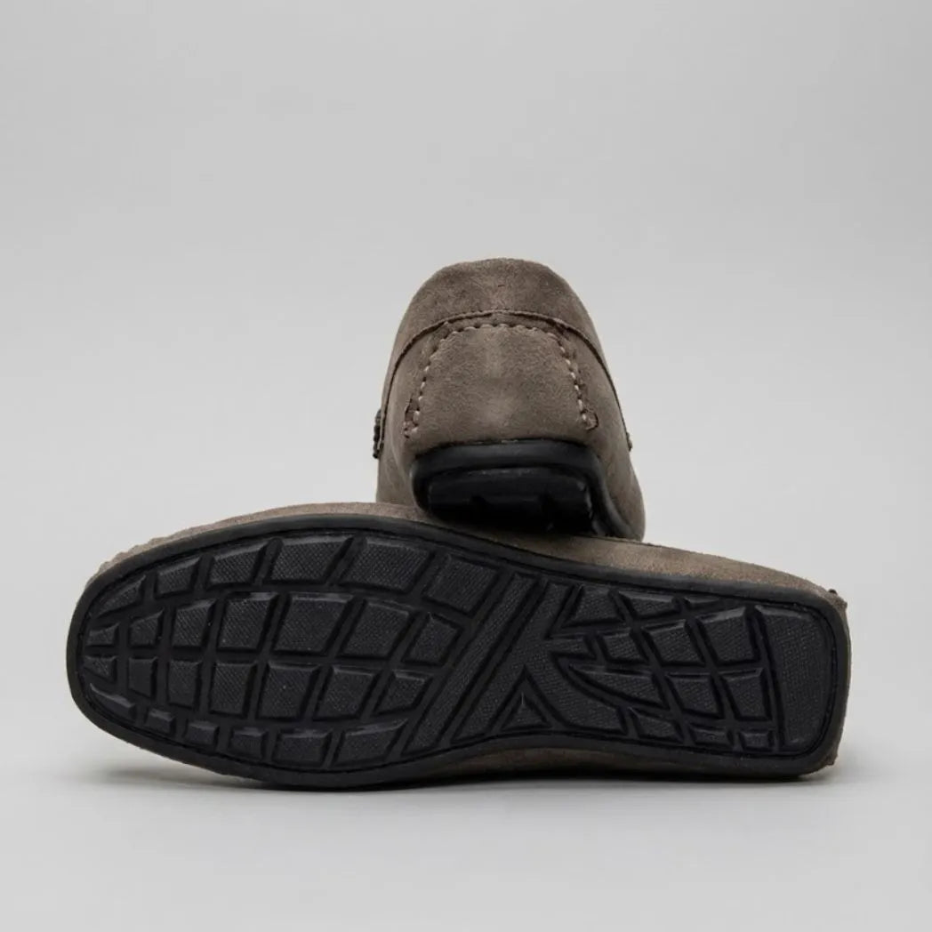 Par do modelo Mocassim Milano Driver com Fivela, na cor cinza-esverdeado, com um dos solados voltado para a câmera.