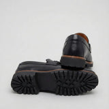 Par do modelo Mocassim Napoli com Fivela, na cor preto, com um dos solados voltado para a câmera.