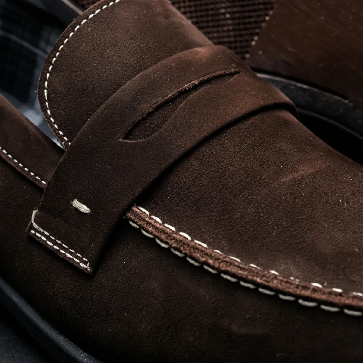 Detalhe do penny loafer Milano Cognac em camurça marrom escura, com faixa lisa e costura clara.