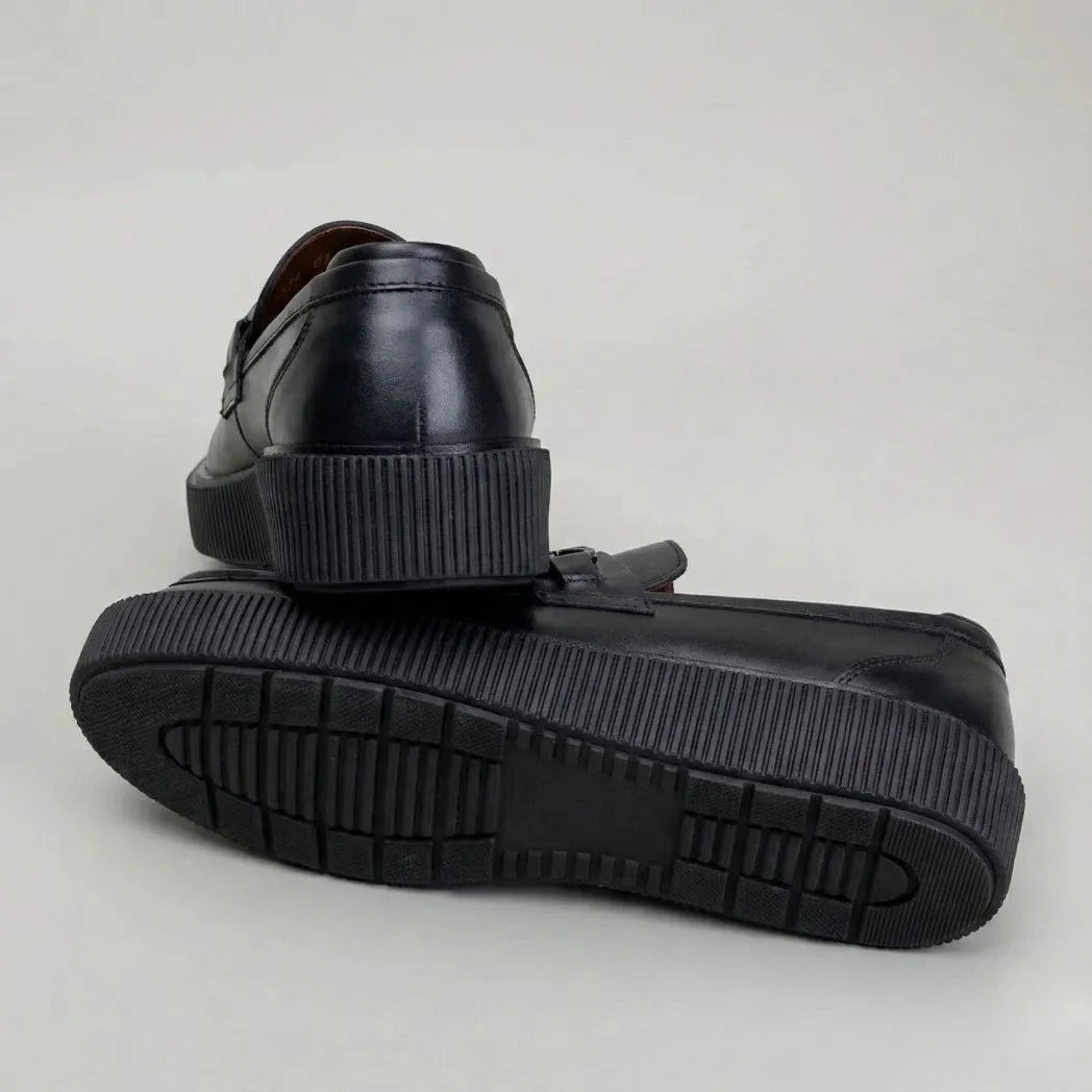 Par do modelo Mocassim Plataform com Fivela, na cor preto, com um dos solados voltado para a câmera.