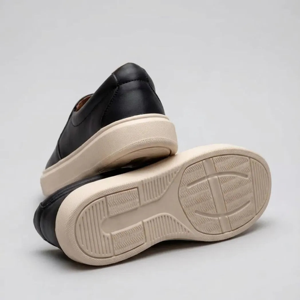 Mocassim Slip On Casual de Couro: par preto com um sapato virado, mostrando a sola bege com sulcos.