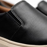 Mocassim Slip On Casual de Couro: detalhe da superfície preta, da costura lateral e da borda da sola bege.