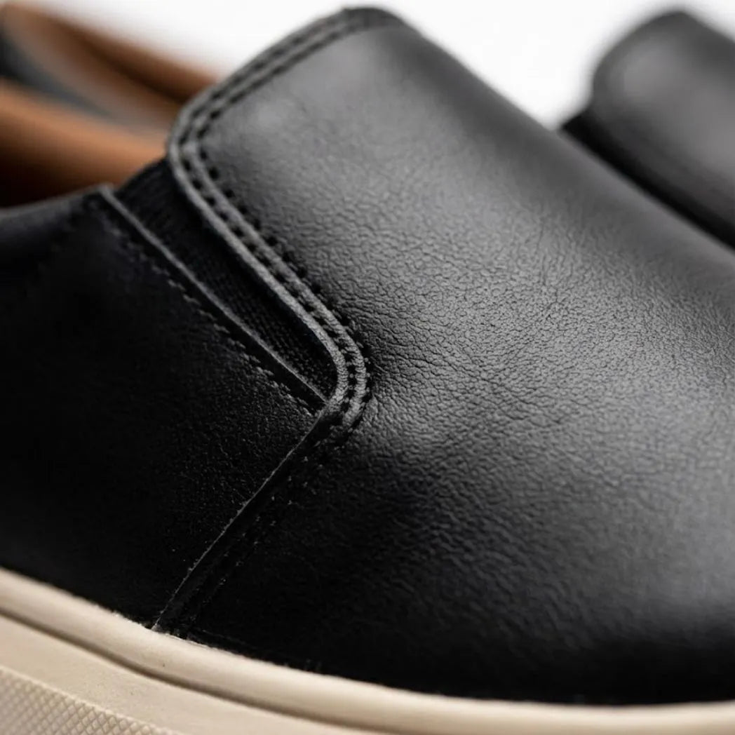 Mocassim Slip On Casual de Couro: detalhe da superfície preta, da costura lateral e da borda da sola bege.