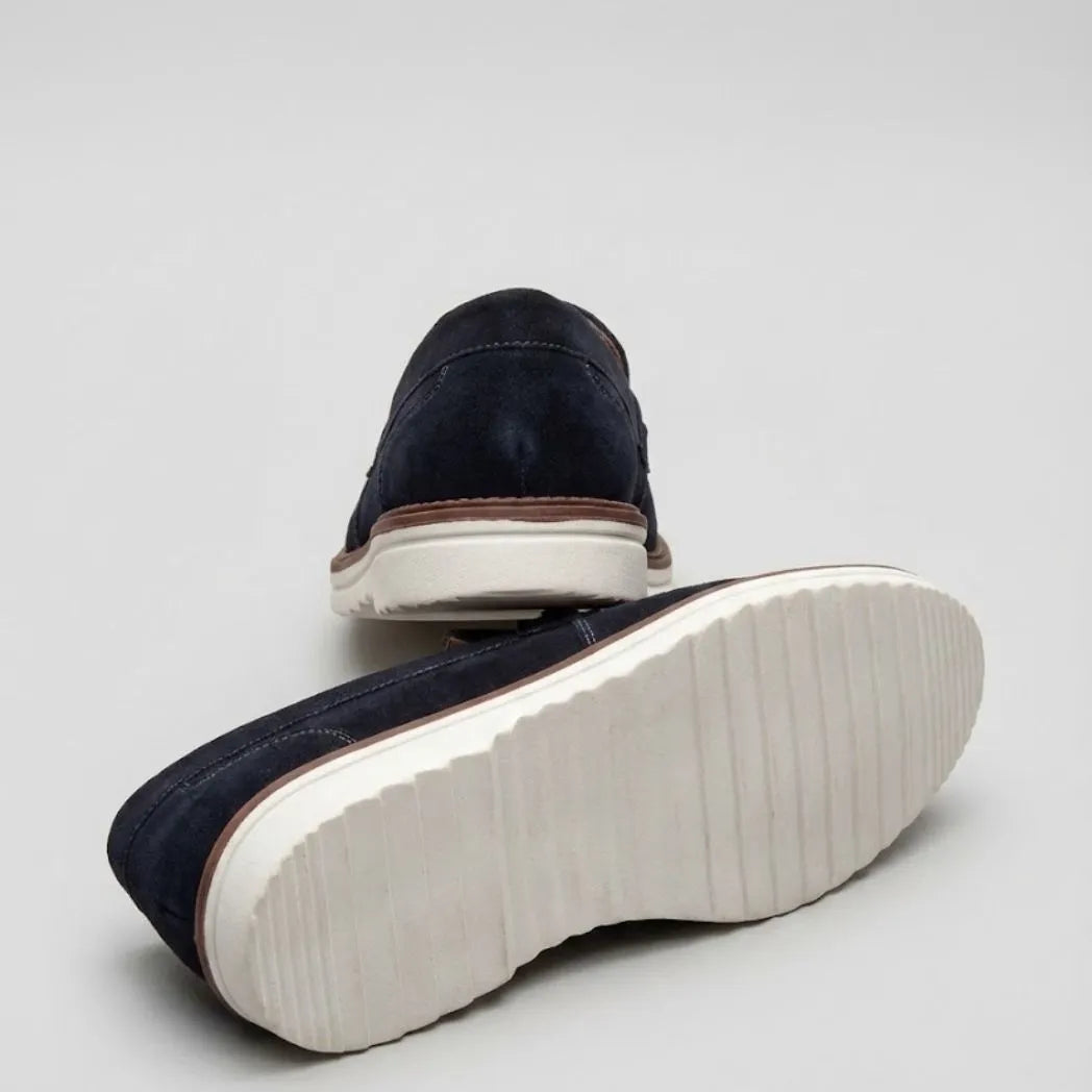 Par do modelo Mocassim Suede Tratorado de Camurça, na cor azul-escuro, com um dos solados voltado para a câmera.