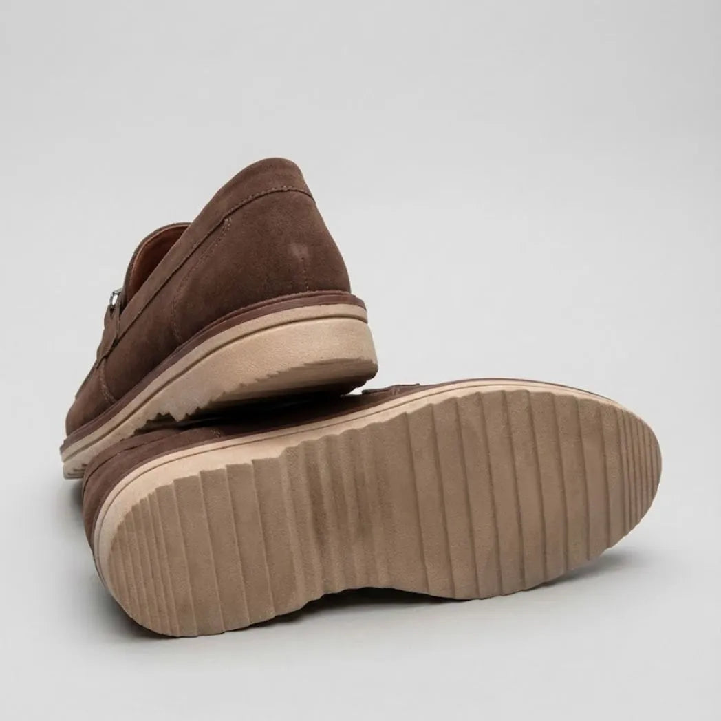 Par do modelo Mocassim Suede Tratorado de Camurça, na cor marrom, com um dos solados voltado para a câmera.