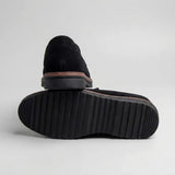 Par do modelo Mocassim Suede Tratorado de Camurça, na cor preto, com um dos solados voltado para a câmera.