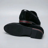 Mocassim Tassel de Camurça Premium: par preto com um sapato virado, mostrando a sola escura e o detalhe vermelho no salto.