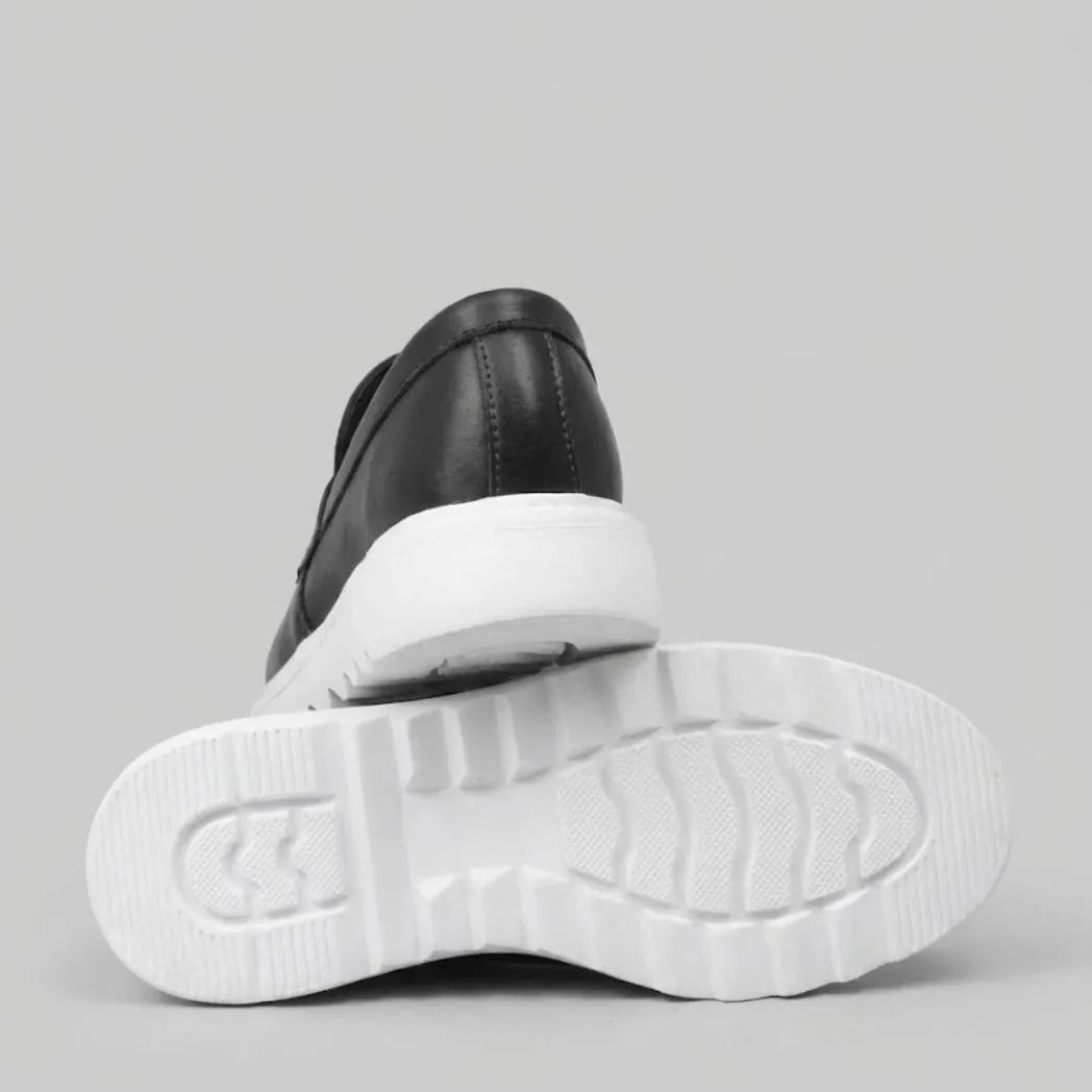 Par do modelo Mocassim Torino Ripple Tratorado de Couro, na cor preto, com um dos solados voltado para a câmera.