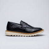 Mocassim Tratorado Siena Ripple: modelo preto de perfil, com faixa frontal, borda marrom e sola bege com sulcos em zigue-zague.