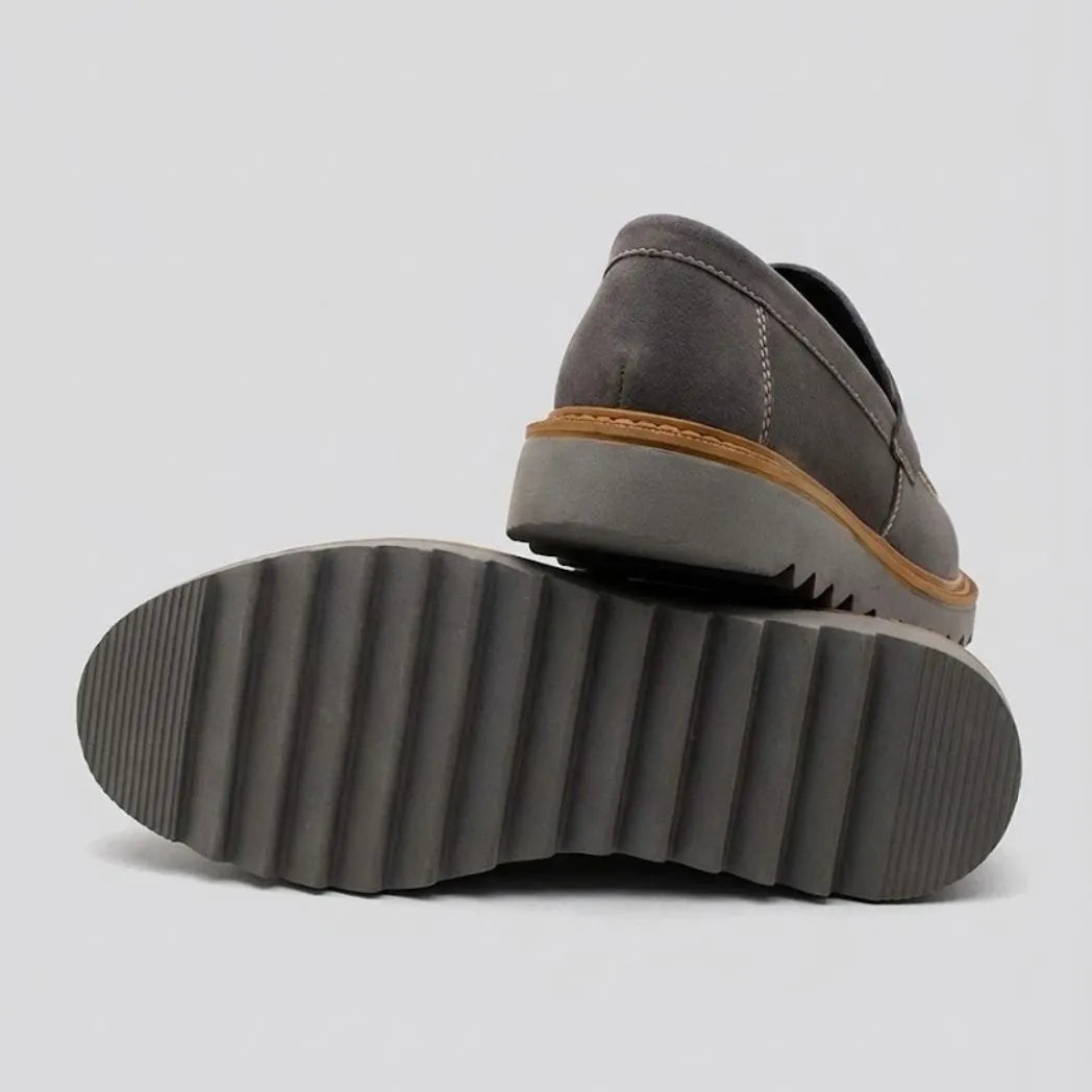 Par do modelo Mocassim Tratorado Taupe de Camurça, na cor cinza, com um dos solados voltado para a câmera.