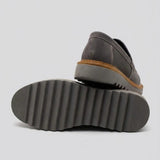 Par do modelo Mocassim Tratorado Taupe de Camurça, na cor cinza, com um dos solados voltado para a câmera.