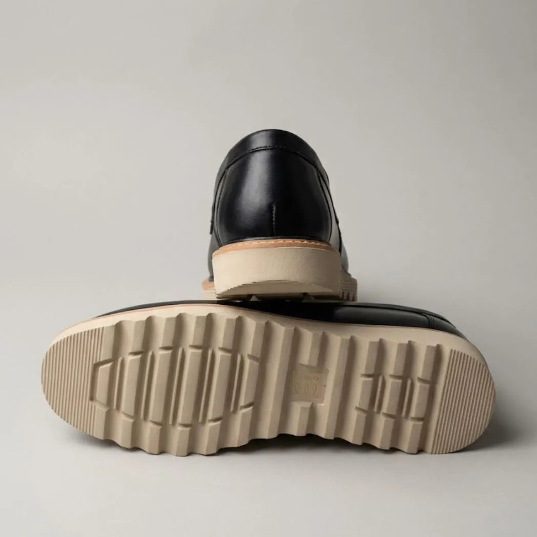 Par do modelo Mocassim Tratorado de Couro Verona, na cor preto, com um dos solados voltado para a câmera.