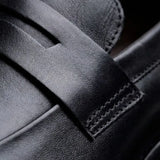 Mocassim Turim Loafer Couro: detalhe da superfície preta, da faixa frontal e da costura lateral.